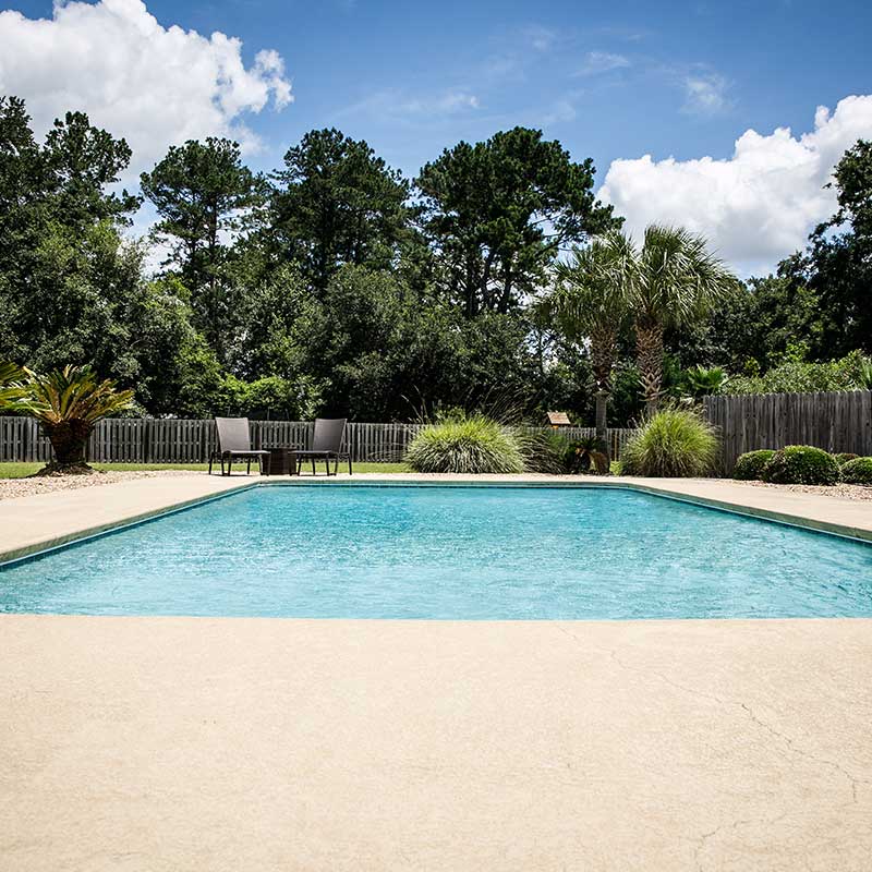 Fenced-in pool area with shrubs, table, and trees in the background.